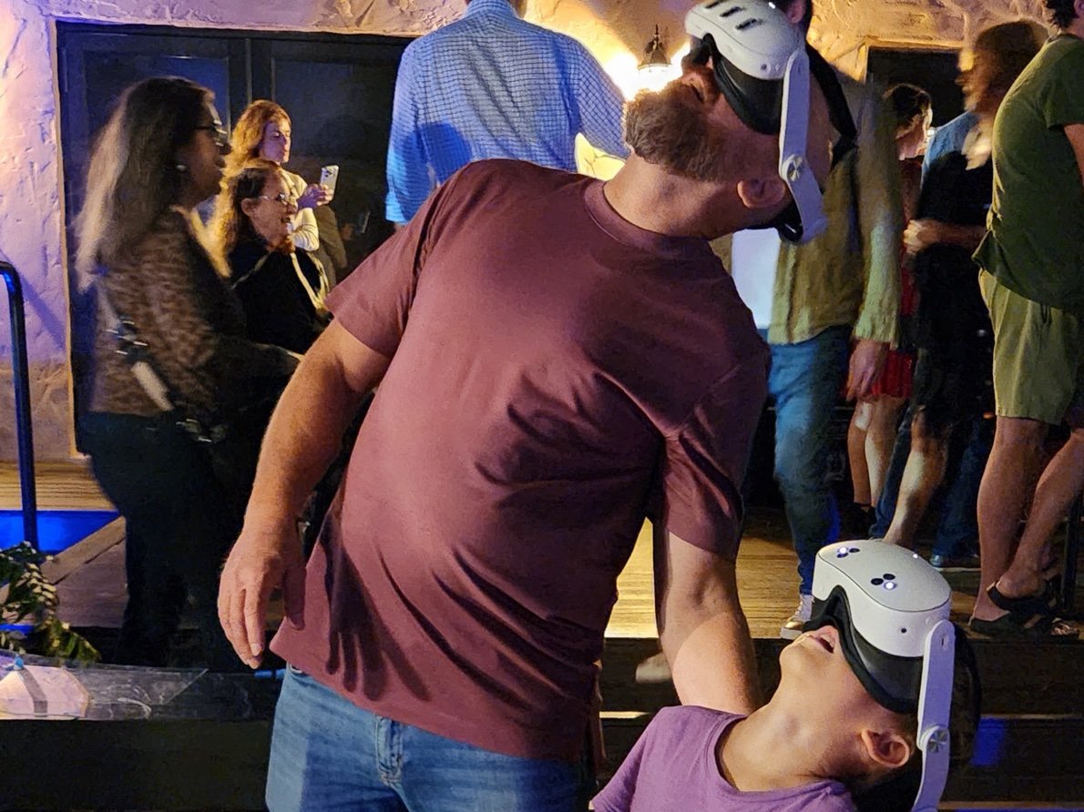 A father and his young son, both in headsets, look up at the same sky